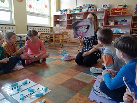 Dzieci siedzą na podłodze i patrzą na ilustrację zębów z próchnicą, którą pokazuje im pani, która klęczy.