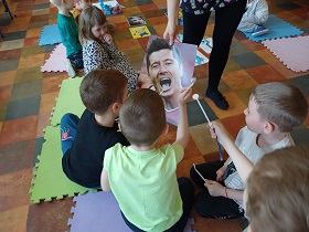 Dzieci siedzą na matach i zaglądają lustereczkami na patyku do zębów na ilustracji, którą pokazuje im pani. Na ilustracji znajduję się Robert Lewandowski. 