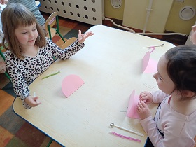 Trzy dziewczynki siedzą przy stole i mają przed sobą różowe karteczkami złożone na pół w półkole. 