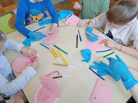 Dzieci siedzą przy stoliku i naklejają białe naklejki bądź rysują coś flamastrami na różowych kartkach, które mają wyglądać jak buzie. Trójka dzieci ma nałożone niebieskie rękawiczki. 