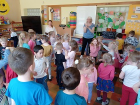 Dzieci stoją w sali w grupce i patrzą na panią, która pokazuje im ruch taneczny. 