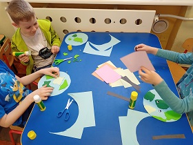 Dzieci siedzą przy stoliku i przyklejają elementy do niebieskiego koła. Na stoliku leżą zielone elementy, kleje, nożyczki oraz kolorowe karteczki i czarne paski. 
