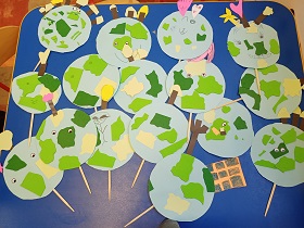 Prace plastyczne dzieci leżą na niebieskim stoliku. Są to papierowe kule ziemskie, z zielonymi kontynentami. Kule ziemskie są przyczepione do drewnianych patyczków. 