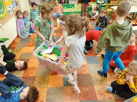Dzieci siedzą w kole na podłodze. Przez środek idą trzy dziewczynki niosące duże pudło z klockami. 