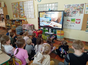 Dzieci siedzą na podłodze i krzesłach przed monitorem, na którym wyświetlany jest film. Widać na nim panią w okularach, która przypatruje się stronie książki.