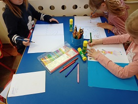 Trzy dziewczynki siedzą przy stoliku i rysują ilustracje na białych kartkach. Na stoliku leży opakowanie flamastrów, kleje i kredki.