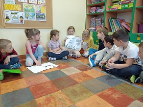 Dzieci siedzą obok siebie i patrzą na dziewczynkę w białej bluzce, która pokazuje im ilustracje, które narysowała. 