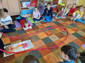 Dzieci siedzą w kole. Przed Panią znajduje się ilustracja przedstawiająca dziewczynkę z zaznaczonymi narządami układu pokarmowego. Wokół tej ilustracji prowadząca rozłożyła czerwoną linę, aby zademonstrować jak długie jest jelito. 