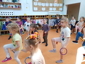 Dzieci ustawione w rozsypce trzymają w dłoni kolorowy okrąg. Część dzieci stoi na jednej nodze i próbuje przełożyć okrę pod podniesioną nogą.