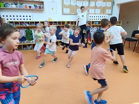 Dzieci biegną trzymając w dłoniach kolorowe okręgi, które imitują kierownicę. Prowadząca trzyma uniesioną rękę w której trzyma zielony okręg.