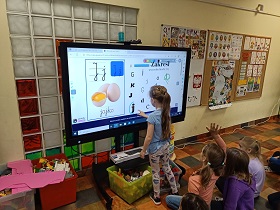 Na ekranie znajduje się ilustracja i napis jajko oraz różne litery pisane różnorodnymi czcionkami. Przed ekranem stoi dziewczynka, która zaznacza odnalezioną wśród innych liter literę j. 
