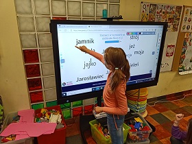 Na ekranie umieszczone są wyrazy zawierające literę j. Przed ekranem stoi dziewczynka, która zaznacza literę j w wyrazie jamnik. 