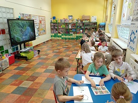 Dzieci siedzą przy stolikach i rysują pastelami po białych kartkach A4. Na monitorze wyświetlany jest film z roślinnością i wodą. 