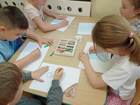 Dzieci siedzą przy stoliku i rysują pastelami po białych kartkach A4. Na środku stołu leży opakowanie pasteli. 