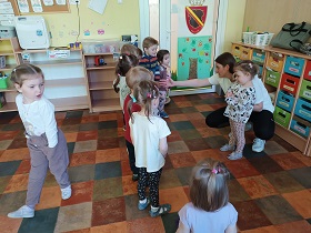 Dzieci stoją przed prowadzącą zajęcia, która kuca i zwraca się do dzieci mówiąc, jak wykonać ćwiczenie.