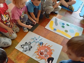 Dzieci siedzą na podłodze, przed nimi leżą dwie plansze. Na pierwszej przedstawione są okrągłe kształty, zaś na drugiej podłużne.