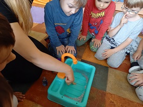 Dzieci siedzą na podłodze, przed nimi stoi plastikowa umywalka napełniona wodą do której pani wkłada palec.