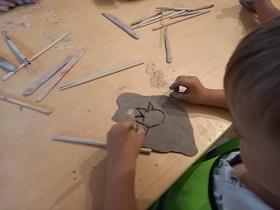 Przy stole stoi chłopiec, przed nim ułożona jest glina w kształcie owalu. Chłopiec w prawej dłoni trzyma patyczek i rysuje uśmiech na glinie.