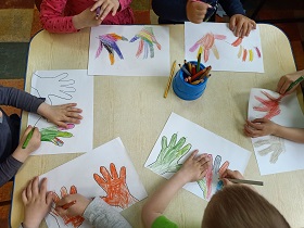 Na stole leży 6 rysunków przedstawiających odrysowane dłonie. Na środku stołu stoi pojemnik z kredkami. Czworo dzieci koloruje swój rysunek.