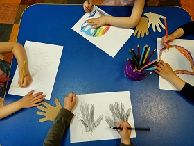Na stole leżą 4 rysunki przedstawiające odrysowane dłonie. Na środku stołu stoi pojemnik z kredkami. Obok rysunków leża szablony odrysowanych dłoni. Dzieci kolorują swoje rysunki. 