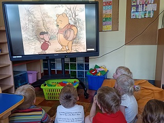 Dzieci siedzą przed tablicą multimedialną i oglądają bajkę pt. Kubuś Puchatek i przyjaciele. Na ekranie widnieje Kubuś Puchatek i Prosiaczek. 
