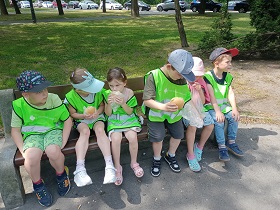 Dzieci w zielonych, odblaskowych kamizelkach siedzą na ławce i jedzą bułki.