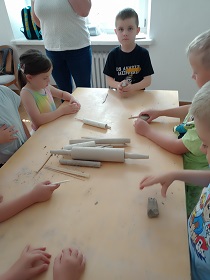 Dzieci pracują przy stole na stojąco. Używają drewnianych przyrządów, tworząc coś z gliny.