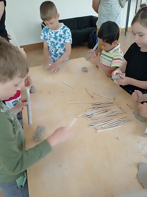 Dzieci pracują przy stole na stojąco. Używają drewnianych przyrządów, tworząc coś z gliny.