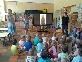 Dzieci siedzą na podłodze przed monitorem, na którym wyświetlany jest film z dziewczynką w czarnej bluzce i spódnicy. Przed dziećmi stoją trzy panie, które są zwrócone do monitora. 