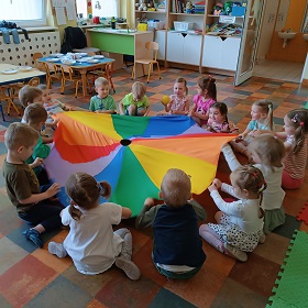 Dzieci siedzą na podłodze wokół kolorowej chusty animacyjnej i falują ją