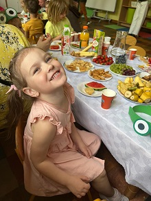 Dziewczynka siedzi przy stole, na którym leżą owoce, ciastka i inne słodkości.