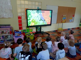 Dzieci siedzą na podłodze przed monitorem, na którym wyświetlana jest bajka. 