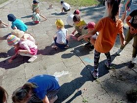 Dzieci kucają lub stoją nad chodnikiem i rysują na nim kolorową kredą. 