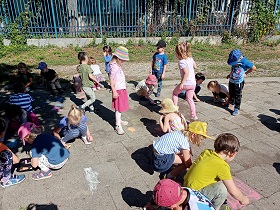 Dzieci kucają lub stoją nad chodnikiem i rysują na nim kolorową kredą. 