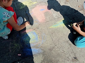 Dwoje dzieci kuca i rysuje kolorową kredą po chodniku. 