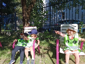 Jest słoneczna pogoda. Dwóch chłopców siedzi na jednym krześle. Obok siedzi dziewczynka, która wskazują na coś palcem. Dzieci ubrane są w kamizelki odblaskowe. 