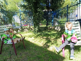 Jest słoneczna pogoda. Po lewej stronie na jednym krześle siedzą 2 uśmiechnięte dziewczynki a po prawej siedzi 1 uśmiechnięta dziewczynka. Ubrane są w kamizelki odblaskowe. 