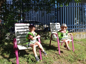 Jest słoneczna pogoda. Chłopiec i dziewczynka siedzą uśmiechnięci na plastikowych krzesłach. Ubrani są w kamizelki odblaskowe. 