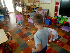 Dzieci biegają po sali i podnoszą małe kartoniki, które leżą na podłodze.