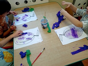 Troje dzieci siedzi przy stole i maluje wodą kartkę, do której przyczepia fioletową lub zieloną bibułę. 