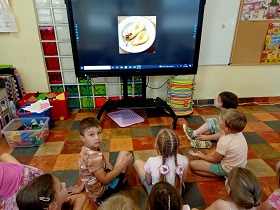 Dzieci siedzą na podłodze przed monitorem, na którym wyświetlane jest zdjęcie deseru z gruszki. 
