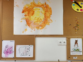 Na korkowej tablicy wiszą 2 pokolorowane przez dzieci obrazki, które przedstawiają ich znaczki. Maria -kuta, Sofia- niedźwiedź. Nad obrazkami wisi duże słońce pomalowane farbami przez dzieci. 