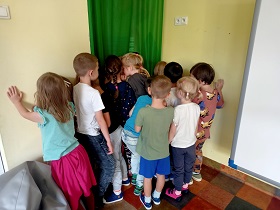 Dzieci stoją w koncie i mają odwrócone głowy w stronę ściany lub zielonej zasłony. 