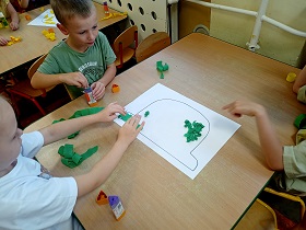 Troje dzieci siedzi przy stoliku i przykleja zieloną bibułę do kartki, na której narysowany jest kubeł. 