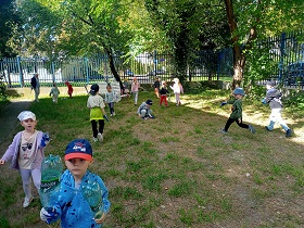 Dzieci biegają lub chodzą po trawie i szukają śmieci. Chłopiec w czapce z czerwonym daszkiem, trzyma dwie plastikowe butelki. 