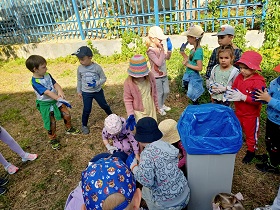 Dzieci segregują śmieci do kosza. Część dzieci stoi obok kosza, a część przy nim kuca.