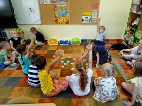 Dzieci siedzą na podłodze i podnoszą swoją rękę do góry. Jedna dziewczynka na środku kółka wybiera sobie kartonik. 