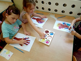 Troje dzieci siedzi przy stole i maluje farbami po kartce, na której znajduję się brązowy pień drzewa. Na stole znajduję się również paleta z farbami. 