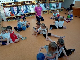 Dzieci w strojach gimnastycznych siedzą w parach na podłodze. Jedna osoba z pary trzyma w rękach piłkę, którą stara się podać drugiej osobie. 