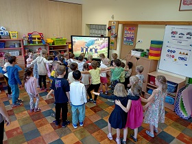 Dzieci stoją przed monitorem i trzymają się za ramiona. Na monitorze wyświetlany jest film, na którym postaci tańczą. 